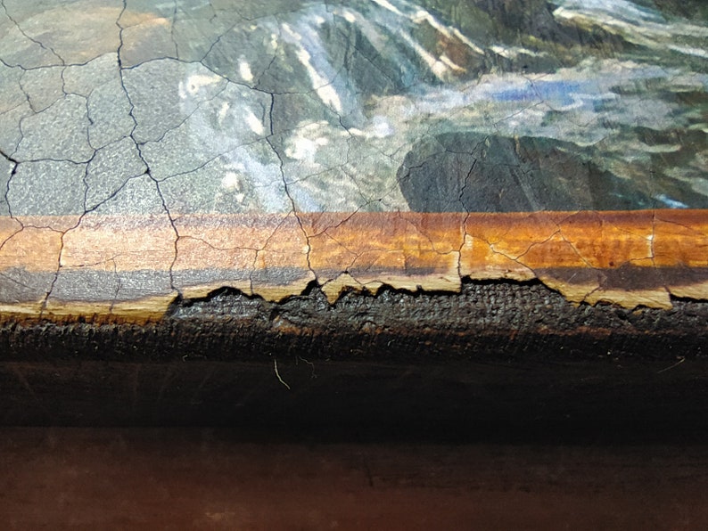 May include: Close-up of a painting with a cracked, brown frame. The painting depicts a landscape with water and rocks. The frame is damaged and peeling.