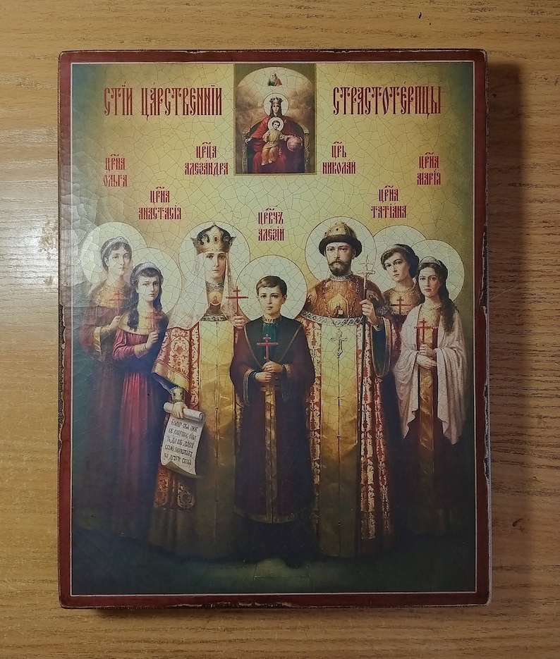 May include: A vintage-style religious icon depicting a family of saints. The icon features a central image of a young boy saint, surrounded by six other saints, including a woman holding a scroll. The icon is framed in a wooden frame with a red border.