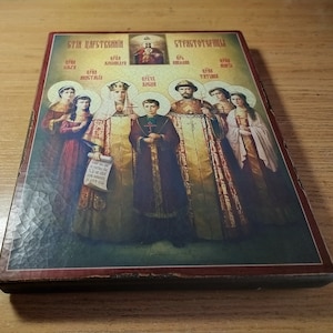 May include: A framed religious icon depicting a family of seven people. The icon is painted in a traditional style with gold accents. The text at the top of the icon reads "Эти царственные" and "Страстотерпцы".