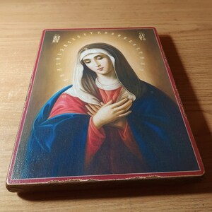 May include: A religious icon depicting the Virgin Mary with a blue and red robe, a white veil, and a halo. The icon is painted on a wooden panel with a red border and features Cyrillic text around the halo.
