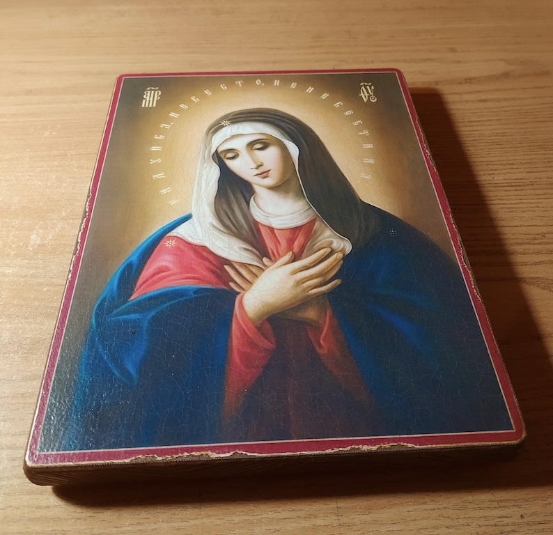 May include: A religious icon depicting the Virgin Mary with a blue and red robe, a white veil, and a halo. The icon is painted on a wooden panel with a red border and features Cyrillic text around the halo.