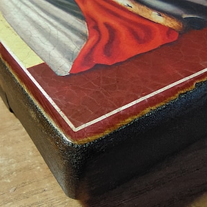 May include: Close-up of the bottom edge of a wooden framed painting. The frame is painted black with a white border around the red painted surface. The painting depicts a religious scene with a figure in a white robe and a red cloak.