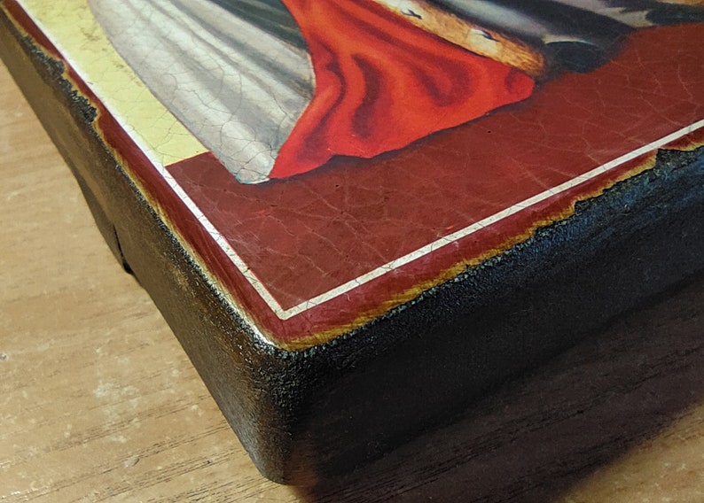 May include: Close-up of the bottom edge of a wooden framed painting. The frame is painted black with a white border around the red painted surface. The painting depicts a religious scene with a figure in a white robe and a red cloak.
