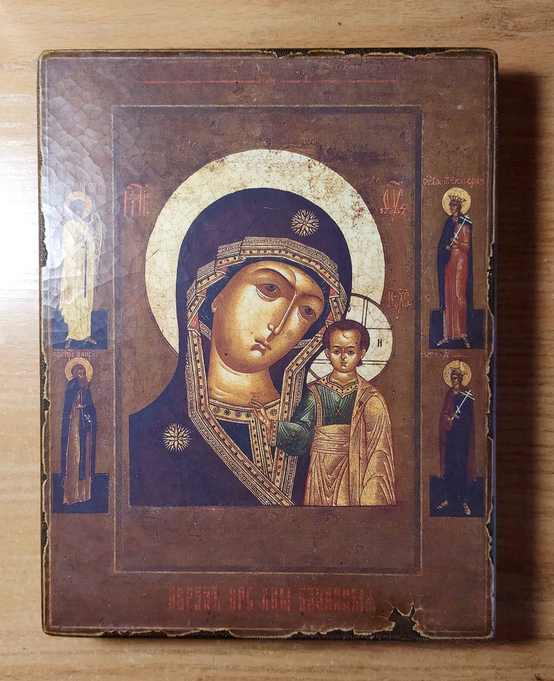 Puede incluir: Un icono religioso de estilo vintage que representa a la Virgen Mar&iacute;a sosteniendo al ni&ntilde;o Jes&uacute;s. El icono est&aacute; enmarcado en un marco de madera oscura y presenta un fondo dorado con detalles intrincados. El texto en la parte inferior dice "ОБОЖЕНИЕ ВСЯКИМ СВЯТЫМ".