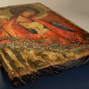 May include: A weathered wooden icon depicting a religious figure with red and blue clothing. The icon is painted on a wooden panel and has a distressed, antique look.