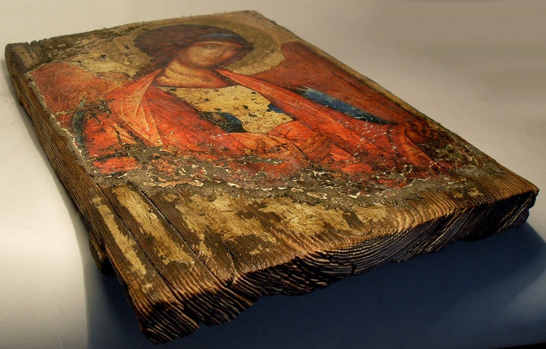 May include: A weathered wooden icon depicting a religious figure with red and blue clothing. The icon is painted on a wooden panel and has a distressed, antique look.