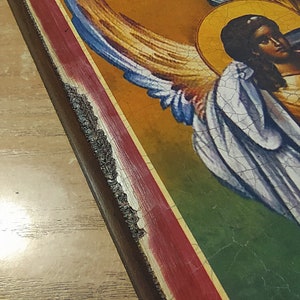 May include: A vintage-style religious icon with a painted image of an angel with wings. The icon is framed in a dark wood frame with a red and gold border.