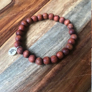 Pode incluir: Um bracelete de contas de madeira com um fecho de prata. As contas são de cor castanho-avermelhado escuro e o fecho tem um desenho de árvore.