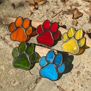 May include: Five stained glass paw prints in different colours: orange, red, yellow, green, and blue. The paw prints are all the same size and shape, and they are arranged in a semi-circle.