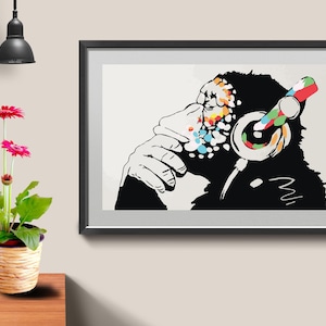 May include: Black and white framed art print featuring a chimpanzee wearing headphones. The chimpanzee's face has colorful paint splatters, and the headphones are decorated with a rainbow pattern. A potted plant with pink flowers is on a wooden table.
