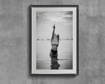 May include: A black and white photograph of a person standing in the water, wearing a white t-shirt pulled over their head and black bikini bottoms. The person is raising their middle finger towards the sky.