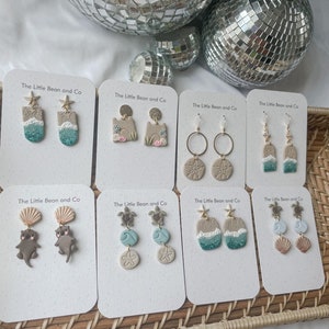 May include: Eight pairs of handmade polymer clay earrings with a beach theme. The earrings feature sea turtles, seashells, starfish, and ocean waves. The earrings are displayed on white cards with the text "The Little Bean and Co."