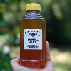 Puede incluir: Una botella de vidrio de miel con una tapa amarilla. La etiqueta dice "Sophia's Honey Raw-Local Honey Annandale, NJ sophiashoney.com". Una ilustración en blanco y negro de una abeja está en la etiqueta.