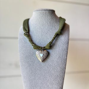 May include: A heart-shaped locket necklace with a green fabric band. The gold-colored locket has an ornate design and a small white heart in the center. The necklace is displayed on a gray jewelry bust.
