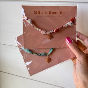 May include: Two fabric necklaces with a gold sun charm pendant. The necklaces are displayed on a pink card with the text "Mila & Rose Co. custom jewelry - charm bar".