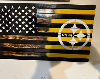 May include: A wooden American flag with a black and yellow Steelers logo on the right side. The flag is made of individual wooden planks and has a distressed look.