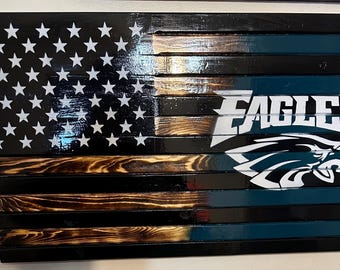 May include: A wooden American flag with a distressed finish. The flag is painted in black, brown, and teal colors. The right side of the flag features the Philadelphia Eagles logo with the word "EAGLES" in white letters.