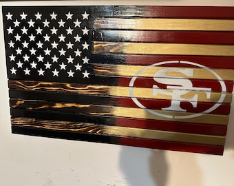 Puede incluir: Una bandera americana de madera con un logotipo de los San Francisco 49ers en el lado derecho. La bandera está hecha de tablones de madera individuales que están teñidos de negro y dorado. El logotipo de los 49ers es blanco sobre un fondo rojo.