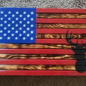 May include: A wooden American flag with a black silhouette of a deer head in the lower right corner. The flag is made of individual wooden planks, and the red stripes are painted red. The blue field of the flag is painted blue, and the stars are white.