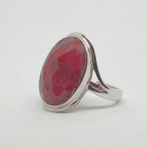 Puede incluir: Un anillo de plata con una gran gema roja ovalada y facetada. La gema es el punto focal, engastada en una banda de plata pulida con un diseño simple y elegante. El anillo se muestra sobre un fondo blanco.