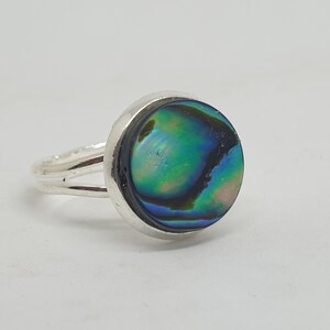 May include: A silver ring featuring a round abalone shell inlay. The shell displays a blue, green, and black iridescent pattern.