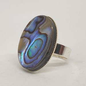 May include: A silver ring featuring an oval abalone shell inlay. The shell displays a blue, green, and brown iridescent pattern.