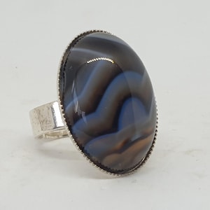 May include: A silver ring with a large, oval stone. The stone has a polished surface with swirling bands of black, blue, and brown. The ring has a simple design. The stone's measurements are approximately 3 cm in length.