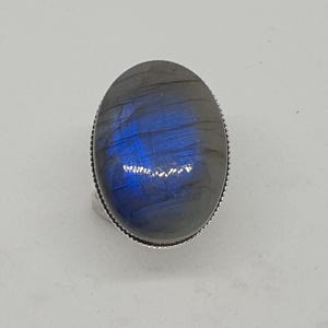 Può includere: Un anello con una grande pietra preziosa di labradorite di forma ovale incastonata in una fascia di metallo argentato. La pietra presenta un colore di base grigio scuro con un evidente riflesso blu iridescente sulla superficie. La montatura ha un bordo.