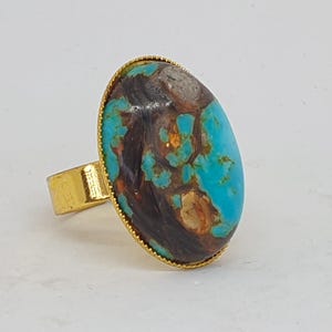 May include: A gold-coloured ring with an oval turquoise stone, displaying brown and black marbling. The stone is set in a gold bezel with a textured edge. The ring has an adjustable band.