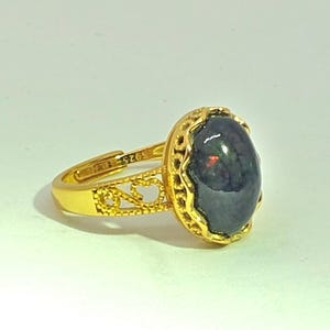 May include: A gold ring with an oval, dark gemstone. The ring has an adjustable band with heart-shaped cutouts and a decorative bezel setting around the stone. The gemstone appears to have iridescent qualities.