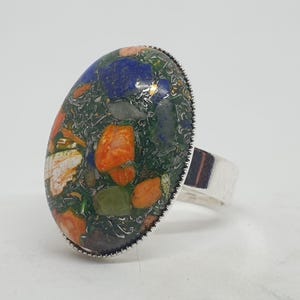 Può includere: Un anello d'argento con una grande pietra ovale incastonata al centro. La pietra è un mix di colori verde, blu, arancione e bianco.