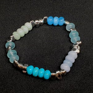 Op de afbeelding: Een armband met turquoise, lichtblauwe en lichtgroene kralen, afgewisseld met zilverkleurige, geometrische kralen. De armband is afgebeeld tegen een donkere achtergrond.