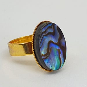 Puede incluir: Un anillo de tono dorado con una incrustación ovalada de concha de abulón. La concha iridiscente muestra patrones de remolinos en tonos azul profundo, morado y verde. El engaste es una banda dorada sencilla con un borde texturizado de cuentas.