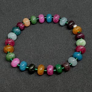Op de afbeelding: Een kleurrijke armband met kralen in verschillende kleuren, waaronder roze, oranje, groen, blauw en paars. De kralen zijn geregen op een groen elastisch koord, wat een levendig en opvallend accessoire creëert.