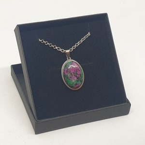 May include: A silver chain necklace with an oval-shaped pendant featuring a green and pink gemstone. The necklace is displayed in a black velvet box.