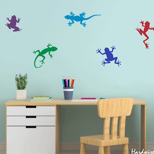 May include: A light blue wall with five colorful animal silhouettes: a purple frog, a blue lizard, a green lizard, a blue frog, and a red frog. A white desk with three drawers and a wooden chair are in front of the wall.