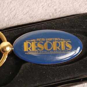 May include: Gold key chain with a blue oval charm that says "Merv Griffin's Resorts Casino Hotel - Atlantic City" in gold lettering.