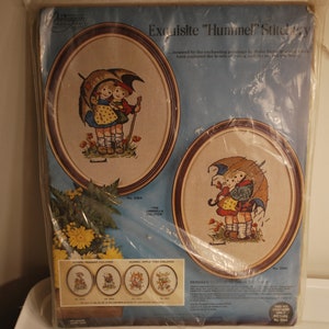 Op de afbeelding: Een vintage naaldpuntset met twee kinderen met paraplu's. De set bevat twee ronde frames met voorgedrukte ontwerpen. De set is gelabeld "Exquisite "Hummel" Stitchery" en bevat het nummer 3082.