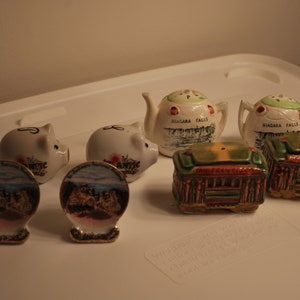 May include: A collection of six vintage ceramic salt and pepper shakers. Two are shaped like teapots with the words "Niagara Falls" on them. Two are shaped like pigs with the words "Atlantic City" on them. Two are shaped like miniature trolleys with green, brown, and red paint.