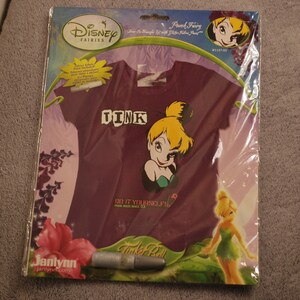 May include: A purple t-shirt with a Tinkerbell design and the word "TINK" on the front. The shirt is packaged with a fabric marker and instructions for "Do It Yourself" customization. The packaging also features a Tinkerbell illustration.