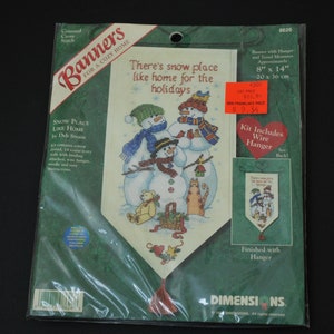 May include: A cross-stitch banner kit featuring a winter scene with three snowmen, a teddy bear, and a cat. The banner is 8 inches by 14 inches and includes a wire hanger. The kit is titled "Snow Place Like Home" by Deb Strain. The text "There's snow place like home for the holidays" is printed on the front of the package.