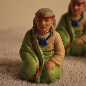 May include: A figurine of a person wearing a green and red robe with a blue and white beaded necklace. The figurine is kneeling with its hands clasped together.