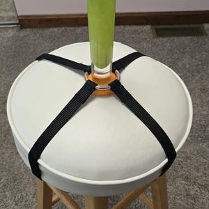 Peut inclure: Un tabouret blanc avec un siège rond et des pieds en bois. Un vase en verre vert est fixé au siège avec des sangles noires et un anneau orange.