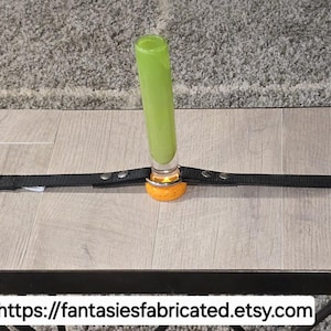 May include: A green glass object is secured to a small table with a black strap. The table has a light wood grain pattern. The base of the glass object is orange. The table is on a gray rug.