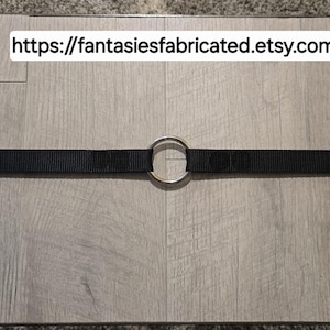 May include: A black nylon strap featuring a centered silver-toned metal O-ring. The strap is horizontally oriented and includes the text https://fantasiesfabricated.etsy.com displayed in a white rectangular box above the center of the strap.