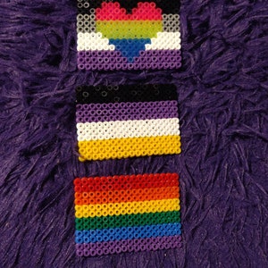 May include: Three small beaded flags, each representing a different sexual orientation. The top flag is purple, white, and black with a rainbow heart in the center. The middle flag is black, purple, white, and yellow. The bottom flag is a rainbow with red, orange, yellow, green, blue, and purple stripes.