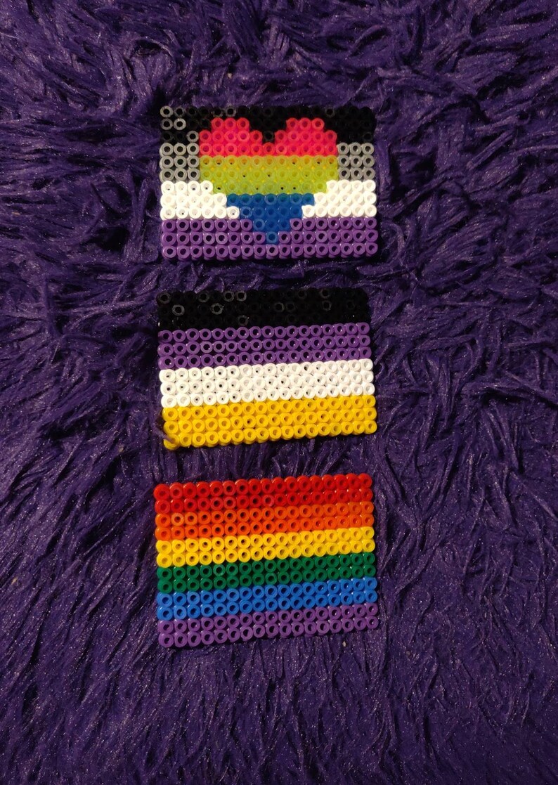 May include: Three small beaded flags, each representing a different sexual orientation. The top flag is purple, white, and black with a rainbow heart in the center. The middle flag is black, purple, white, and yellow. The bottom flag is a rainbow with red, orange, yellow, green, blue, and purple stripes.