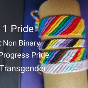 Könnte beinhalten: Vier bunte Freundschaftsbänder, die auf einem braunen Pappröhrchen gestapelt sind. Die Armbänder sind aus Fäden in verschiedenen Farben hergestellt und repräsentieren verschiedene Pride-Flaggen. Die Armbänder sind mit 1 Pride, 2 Non Binary, 3 Progress Pride und 4 Transgender beschriftet.