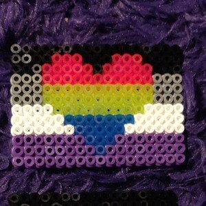 May include: A heart-shaped pride flag made of colorful beads. The flag is made of black, red, yellow, green, blue, white, and purple beads. The heart is in the center of the flag and is made of red, yellow, green, and blue beads.