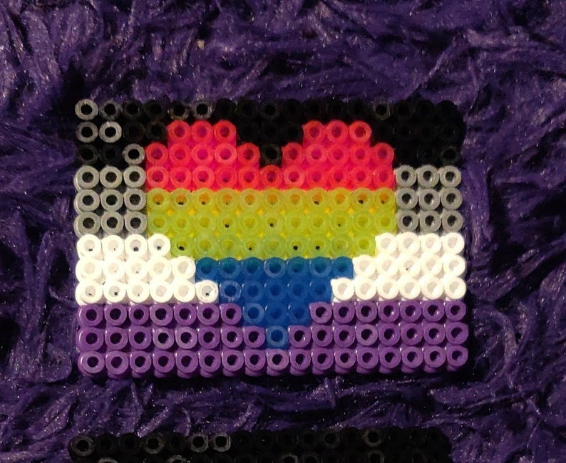 May include: A heart-shaped pride flag made of colorful beads. The flag is made of black, red, yellow, green, blue, white, and purple beads. The heart is in the center of the flag and is made of red, yellow, green, and blue beads.